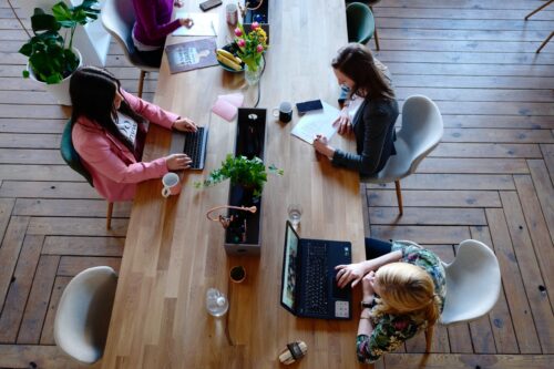 Pracownicy przy stole biurowym, piszą i korzystają z laptopów.