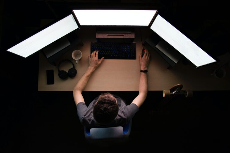Osoba przy biurku z trzema monitorami i klawiaturą.