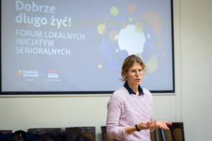 Kobieta przemawiająca na forum inicjatyw senioralnych.